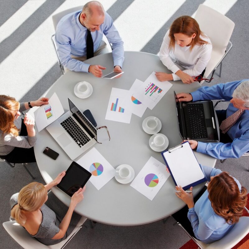 A group of salesmen sit around a table in a sun soaked meeting room.